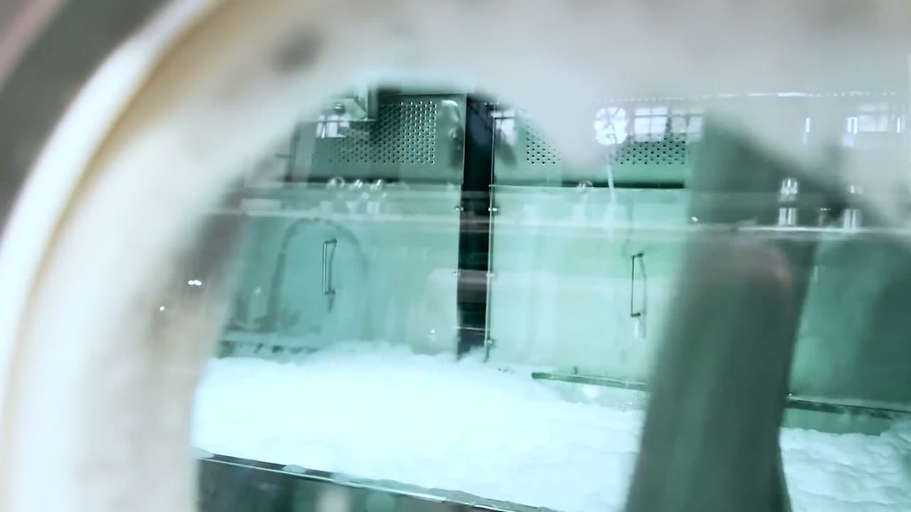 View through the glass door of a commercial washing machine into a laundry, with detergent foam banked against the glass and stainless steel equipment behind it.