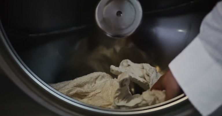 A hand in a white sleeve lowering a cream-coloured towel into the drum of a machine, one item at a time.