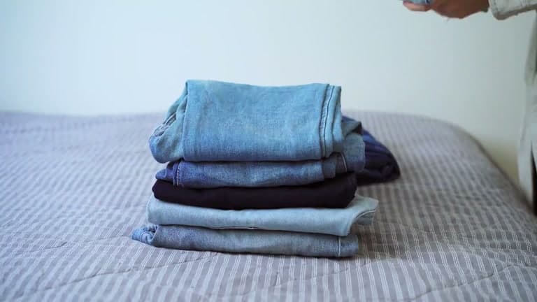 A neat stack of five pairs of folded jeans on a bed, with a hand reaching towards the pile.