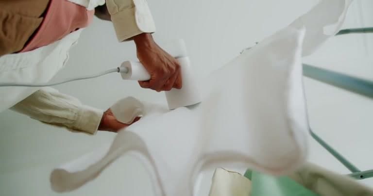 Two hands guiding a steam iron across a white garment spread over an ironing board, seen from below.