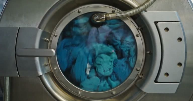 Teal garments tumbling behind the round glass door of a stainless steel commercial cleaning machine.
